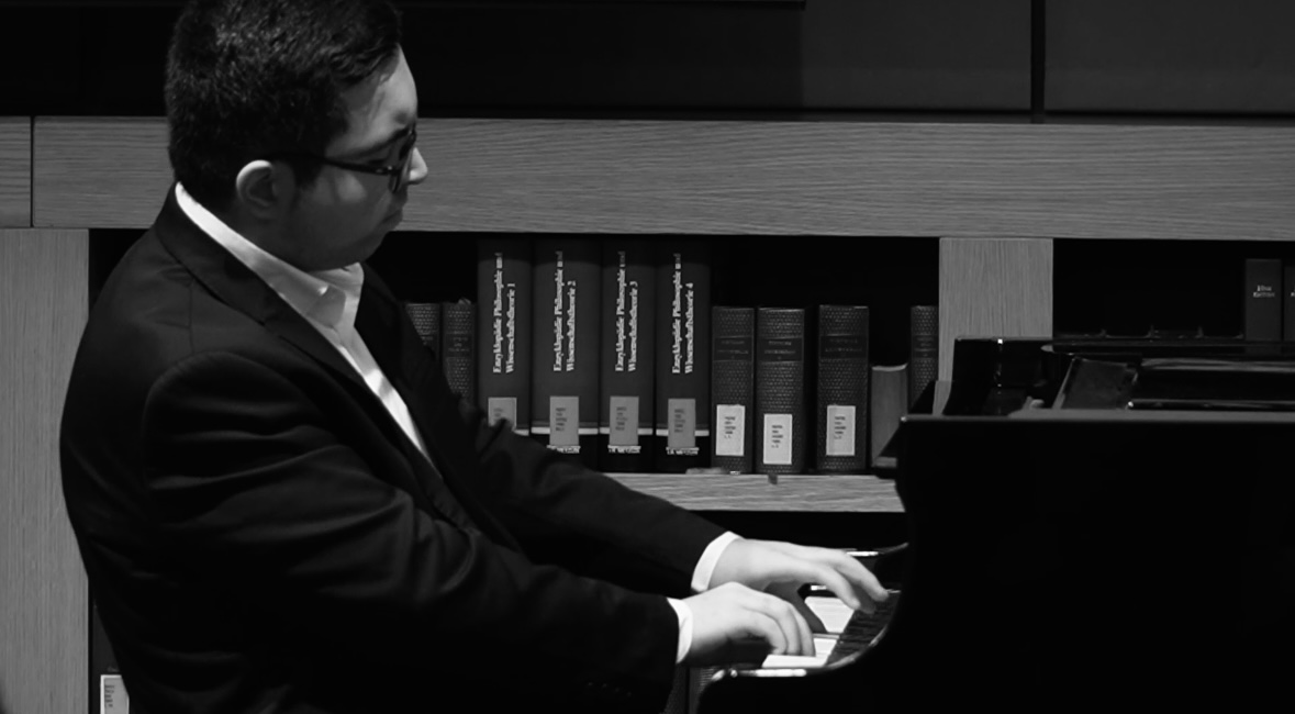 Lefteris Gavalas playing the piano in profile, bookshelves behind him